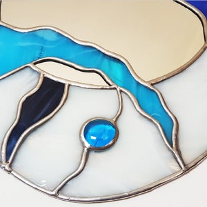 Op de afbeelding: Een ronde spiegel van gekleurd glas met een blauw, wit en zwart ontwerp. De spiegel heeft een zilveren frame en een klein blauw glazen accent.