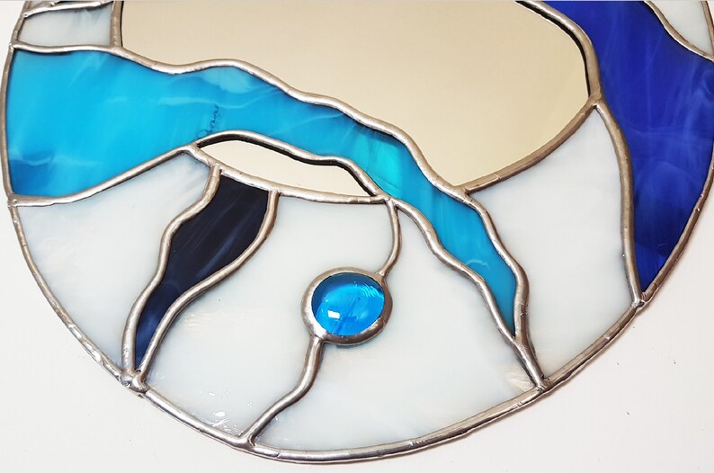 Op de afbeelding: Een ronde spiegel van gekleurd glas met een blauw, wit en zwart ontwerp. De spiegel heeft een zilveren frame en een klein blauw glazen accent.
