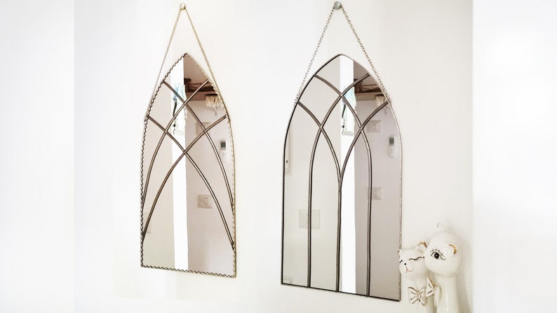 May include: Two arched mirrors with decorative metal frames hang on a white wall. The mirrors are identical in shape and size, but one has a silver frame and the other has a gold frame.