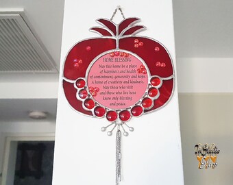 May include: A stained glass pomegranate with red and pink glass pieces. The pomegranate has a silver chain hanging from the bottom. The text on the pomegranate reads: "HOME BLESSING May this home be a place of happiness and health of contentment, generosity and hope. A home of creativity and kindness. May those who visit and those who live here know only blessing and peace."