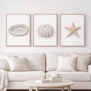 May include: Three framed prints of coastal elements: a shell, a sea urchin, and a starfish. The prints are in neutral tones and hang above a white sofa with matching pillows. A round coffee table and side table complete the living room setting.