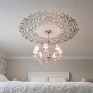 May include: A multi-armed chandelier with pink fabric shades and clear crystal accents hangs from a ceiling featuring a large, circular, intricate gray stencil pattern. The light fixture is centered beneath the decorative ceiling medallion.