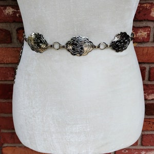 以下が含まれることがあります： 楕円形の金属製チャームが付いたシルバーチェーンベルト。各チャームには、矢印やその他のシンボルが描かれた装飾的なデザインが施されています。