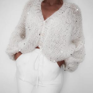 Può includere: Un cardigan bianco lavorato a maglia con scollo a V e chiusura con bottoni. Il cardigan è decorato con paillettes argentate.