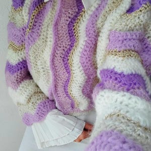 May include: A handmade crochet sweater in shades of lavender, white, and gold. The sweater features a striped pattern with wavy lines and puff sleeves. The sleeves are long and the collar is white.