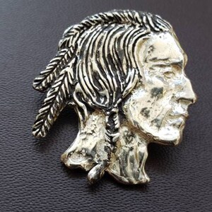 May include: Silver-toned metal pin featuring a detailed profile of a Native American man with a feathered headdress.