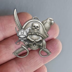 Könnte beinhalten: Silberfarbener Piratenanhänger mit Totenkopf und gekreuzten Knochen, einem Papagei, einem Piratenhut und gekreuzten Schwertern. Der Anhänger hat oben eine Schlaufe zum Aufhängen und einen kleinen Totenkopf auf einem kleineren Hut.