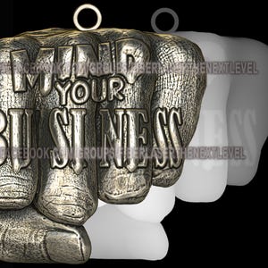 Op de afbeelding: Een zilveren vuistbedel met de woorden "Mind Your Business" erop gegraveerd. De bedel heeft een lus aan de bovenkant om op te hangen.