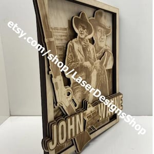 May include: A layered wooden artwork featuring a portrait of John Wayne, a rifle, and the text "JOHN WAYNE". The artwork is made of light brown wood with a dark brown frame and is a decorative piece.
