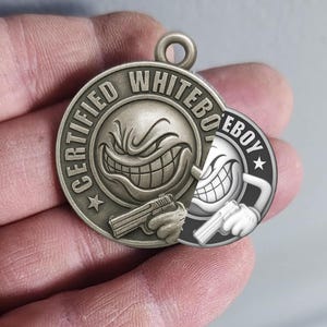 Op de afbeelding: Zilverkleurige hanger met de tekst "CERTIFIED WHITEBOY" rond een grijnzend gezicht met een pistool. Een kleinere, zwart-witte versie van het ontwerp staat rechts. De hanger heeft een lus aan de bovenkant om aan een ketting te bevestigen.