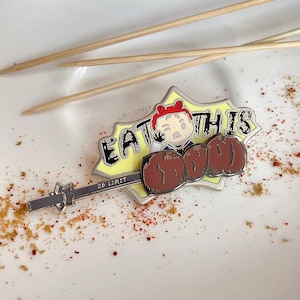 May include: A silver enamel pin featuring a cartoon character holding a skewer of meat with the text "EAT THIS" in a yellow speech bubble. The pin is on a white surface with red and brown spices.
