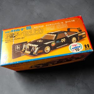 May include: A rectangular cardboard box for a 1/16 scale radio-controlled Dome Celica model car. The box is yellow and red with a black race car graphic, the number 99, and the text Renoma, Dunlop, and Tomy printed on the front.