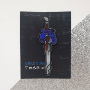 Peut inclure: Un charm en acrylique représentant une épée stylisée avec des détails rouges, argentés et bleus, ornée de roses bleues. Le charm est posé sur un fond noir avec le texte "Devil May Cry" et "Acrylic Charm".
