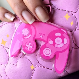 Op de afbeelding: Roze GameCube-controller sleutelhanger met de tekst "Play Me" aan de voorkant. De sleutelhanger is gemaakt van helder plastic met een roze ontwerp.