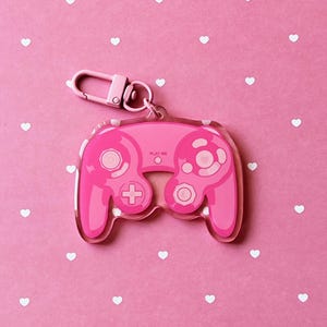Puede incluir: Un llavero rosa de acrílico con forma de mando de videojuegos. El llavero tiene un cierre plateado y un anillo rosa. El mando tiene botones y una cruceta. El fondo es rosa con formas de corazón blancas.