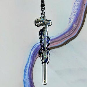 Peut inclure: Un pendentif transparent et irisé avec un design stylisé et allongé, suspendu à une chaîne noire. Le pendentif présente un sommet en forme de croix et un corps tourbillonnant et texturé avec des touches de bleu et d'argent. Le fond présente des coups de pinceau violets et bleus abstraits.