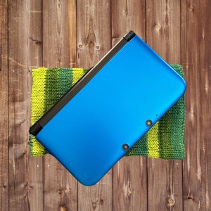 May include: A blue handheld gaming console with a black trim, resting on a green and yellow striped knitted fabric.
