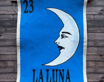 Puede incluir: Una pancarta de lona azul con una ilustración de luna creciente blanca y el número 23. El texto "LA LUNA" está impreso en negro debajo de la luna. La pancarta está sujeta por varillas de madera y clips negros.