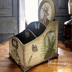 May include: A decorative wooden desk organizer with a vintage aesthetic. Features compartments, a deer portrait, butterfly and fern illustrations, and ornate metal accents. The organizer has a distressed finish and a rectangular shape.