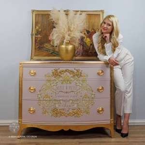 May include: A light pink dresser with gold accents and three drawers. The dresser has a curved bottom and a decorative design on the front with the text "Petit a Petit L'oiseau Fait Son Nid".
