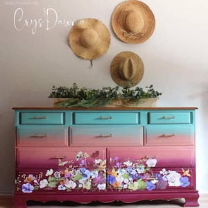 May include: A vintage dresser with a teal, pink, and burgundy ombre paint finish. The bottom drawers feature a floral design. Three straw hats hang on the wall behind the dresser.