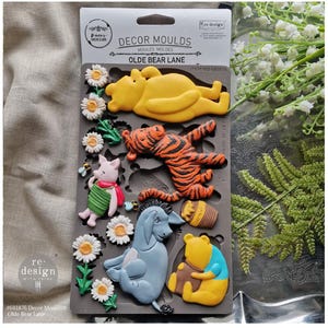 May include: A gray silicone mold featuring Winnie the Pooh characters, including Pooh Bear, Tigger, Piglet, Eeyore, and Pooh with a honey pot. The mold also includes daisy and bee designs. The packaging reads "DECOR MOULDS OLDE BEAR LANE".