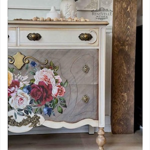 May include: A white dresser with floral decals and gold hardware. The dresser has four drawers and sits on four legs. The dresser is decorated with a floral design featuring roses, leaves, and other flowers. The dresser is painted in a white color with a distressed finish. The dresser is a vintage style piece of furniture.
