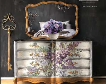 May include: A vintage-style dresser with six drawers, painted in a light gray with a floral design featuring purple and green flowers. The dresser has gold accents and ornate legs. The dresser is in front of a black wall with a gold-framed mirror reflecting a bedroom.