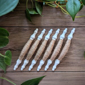 Può includere: Sei bacchette decorative bianche e beige con un involucro di spago naturale. Le bacchette hanno una parte superiore e inferiore bianche bulbose e sono disposte diagonalmente su una superficie di legno. Foglie e viti verdi sullo sfondo.