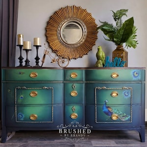 May include: A teal and black dresser with six drawers, featuring a peacock design and gold accents. The dresser has a curved front and sits on four legs. The dresser is decorated with a gold sunburst mirror and candlesticks.