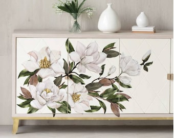 May include: White cabinet with three doors featuring a floral design of white magnolia blossoms and green leaves. The cabinet has gold accents and sits on a white surface.