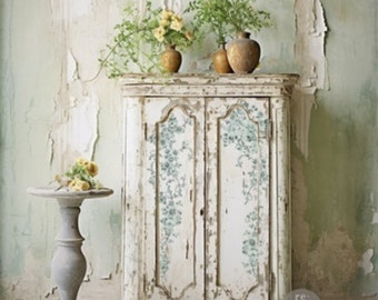 May include: A white antique cabinet with two doors, featuring a floral design. The cabinet is distressed and has a rustic charm. It is decorated with two vases, one with green ferns and the other with yellow flowers.