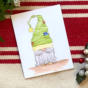Könnte beinhalten: Aquarellillustration eines skurrilen Gnomus mit einem grünen Hut mit gelbem Bommel und Flicken. Der Wichtel hat einen langen, grauen Bart und steht auf einer braunen Oberfläche. Die Karte befindet sich auf einem rot-weiß gestreiften Hintergrund.