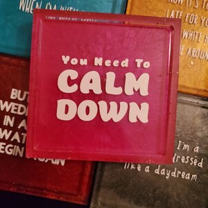 May include: Pink square coaster with the text "You Need To Calm Down" in white letters. The coaster has a glittery finish.