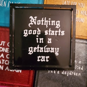 May include: Black square coaster with white text that reads "Nothing good starts in a getaway car".