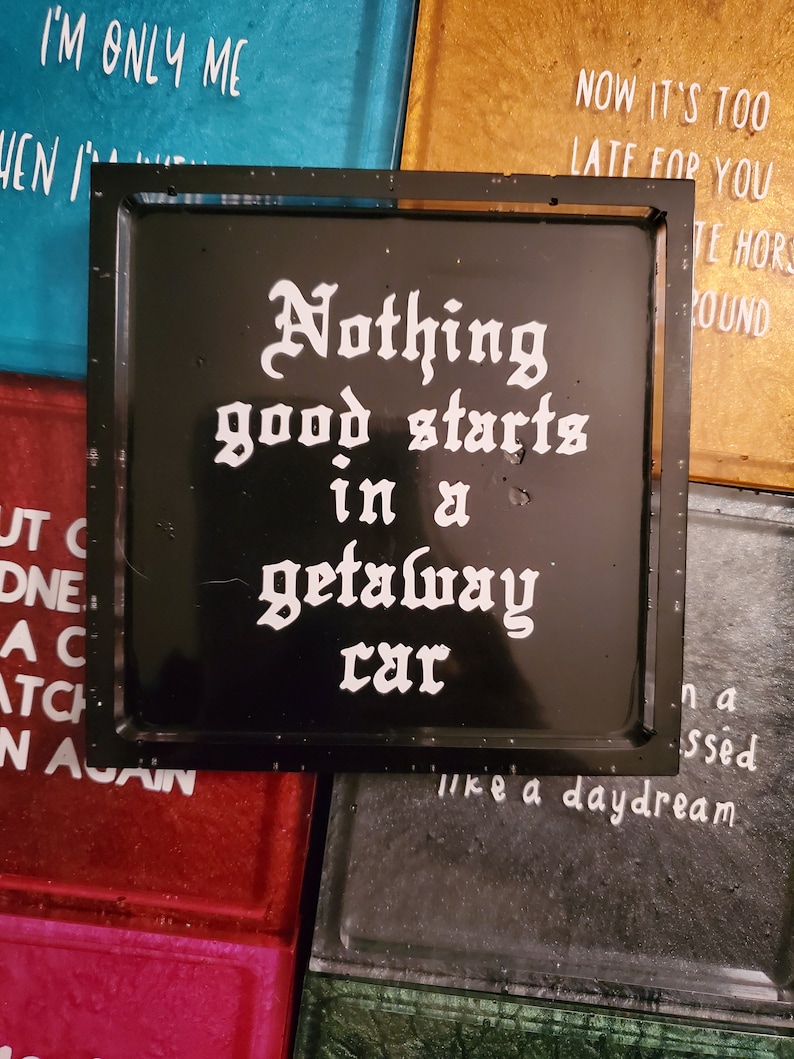 May include: Black square coaster with white text that reads "Nothing good starts in a getaway car".