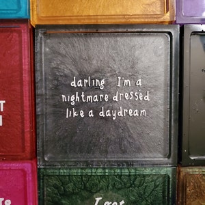 May include: Three square glass tiles with black frames. The tiles have a glittery background and white text. The top tile says "TO COME AROUND". The middle tile says "darling I'm a nightmare dressed like a daydream". The bottom tile says "I got wasted like".
