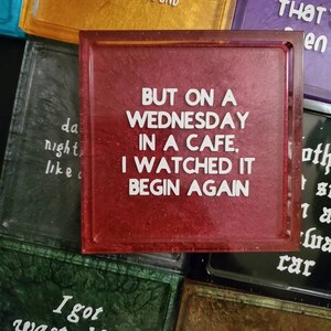 May include: A set of six red, gold, green, purple, black, and brown square coasters with white text. The text on the red coaster reads "But on a Wednesday in a cafe, I watched it begin again."