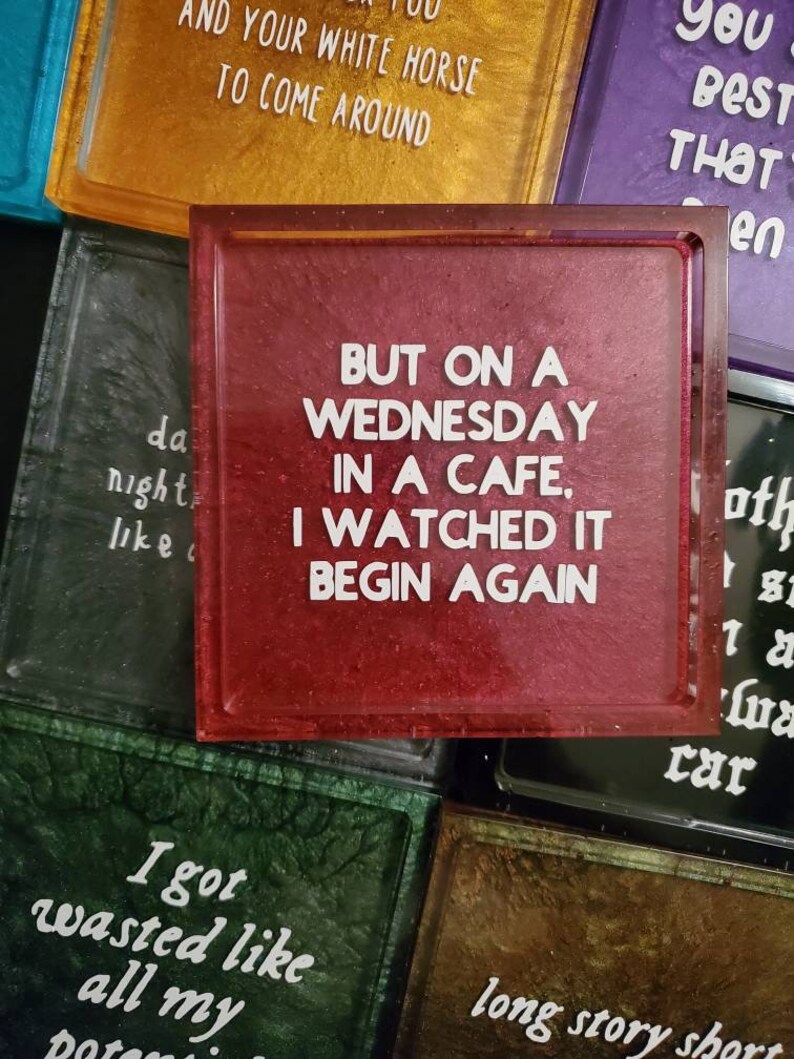 May include: A set of six red, gold, green, purple, black, and brown square coasters with white text. The text on the red coaster reads "But on a Wednesday in a cafe, I watched it begin again."