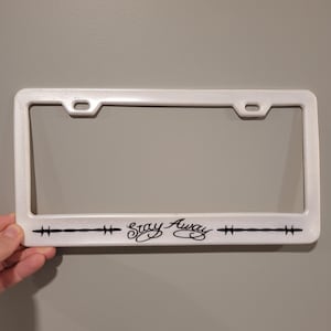 Könnte beinhalten: Weißer Kennzeichenrahmen mit dem Schriftzug "Stay Away" in schwarzer Schrift. Der Rahmen hat ein schlichtes Design mit zwei horizontalen Linien und kreuzartigen Symbolen auf beiden Seiten des Textes.