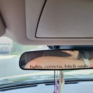 May include: A rearview mirror in a car with a sticker that reads "lights, camera, bitch smile".
