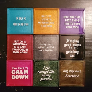 May include: Nine colorful square coasters with white text. The coasters feature various quotes, including "I'm only me when I'm with you", "Now it's too late for you", "You are the best thing that's ever been mine", "But on a Wednesday in a cafe, I watched it begin again", "Darling I'm a nightmare dressed like a daydream", "Nothing good starts in a getaway car", "You need to calm down", "I got wasted like all my potential", and "Long story short, I survived."