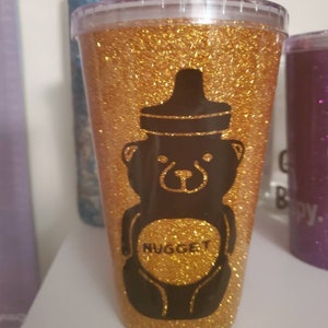 以下が含まれることがあります： 金色のグリッタータンブラーに、黒いクマのシルエットと、お腹に「NUGGET」の文字が書かれています。クマは哺乳瓶をしています。