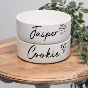May include: Two white ceramic pet bowls stacked. The top bowl has the name "Jasper" in black script with a paw print. The bottom bowl has the name "Cookie" with a heart symbol. The bowls are on a wooden surface.
