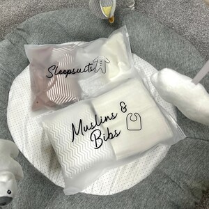May include: Two clear plastic bags labeled "Sleepsuits" and "Muslins & Bibs" rest on a gray and white play mat. The sleepsuit bag contains striped fabric, while the muslin bag holds white items. A baby bib icon is visible.