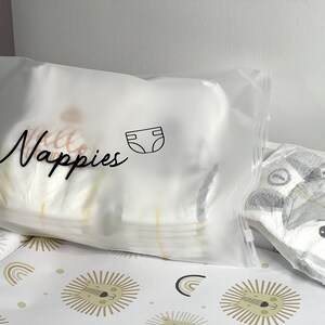 May include: A clear plastic bag filled with diapers, labeled "Nappies" in cursive script, with a diaper icon. A diaper with a fox design is visible. The bag and diaper are on a changing pad with a lion and rainbow pattern.