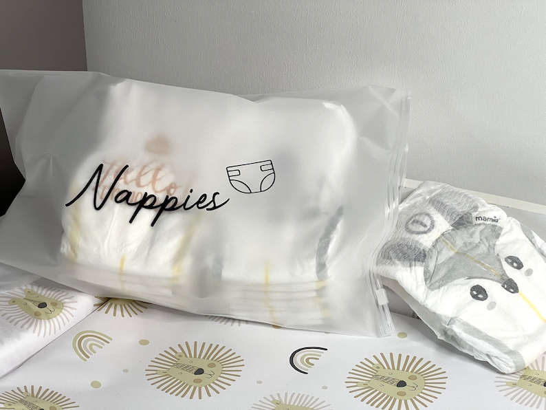May include: A clear plastic bag filled with diapers, labeled "Nappies" in cursive script, with a diaper icon. A diaper with a fox design is visible. The bag and diaper are on a changing pad with a lion and rainbow pattern.