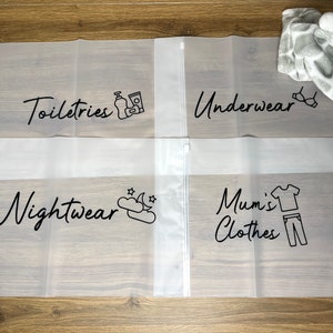May include: Four clear, zippered storage bags with black text and illustrations. The bags are labeled "Toiletries," "Underwear," "Nightwear," and "Mum's Clothes." A plush giraffe toy and a gray blanket are in the background.