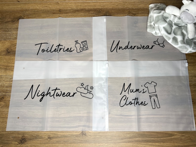 May include: Four clear, zippered storage bags with black text and illustrations. The bags are labeled "Toiletries," "Underwear," "Nightwear," and "Mum's Clothes." A plush giraffe toy and a gray blanket are in the background.