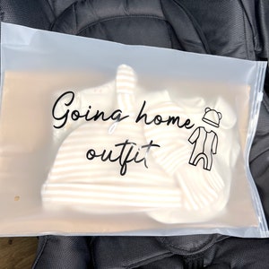 May include: A clear plastic bag with the words "Going home outfit" in black script. Inside the bag are baby clothes, including a striped hat and onesie. A stroller is in the background.